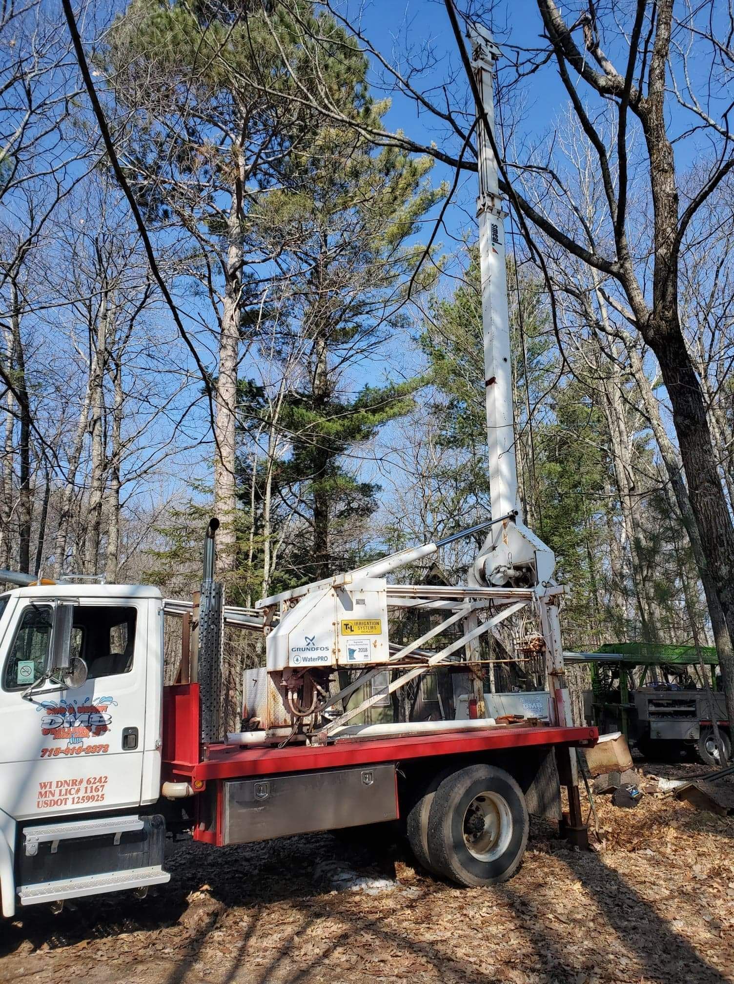 Well drilling truck in a wooded area with a raised white drill mast.