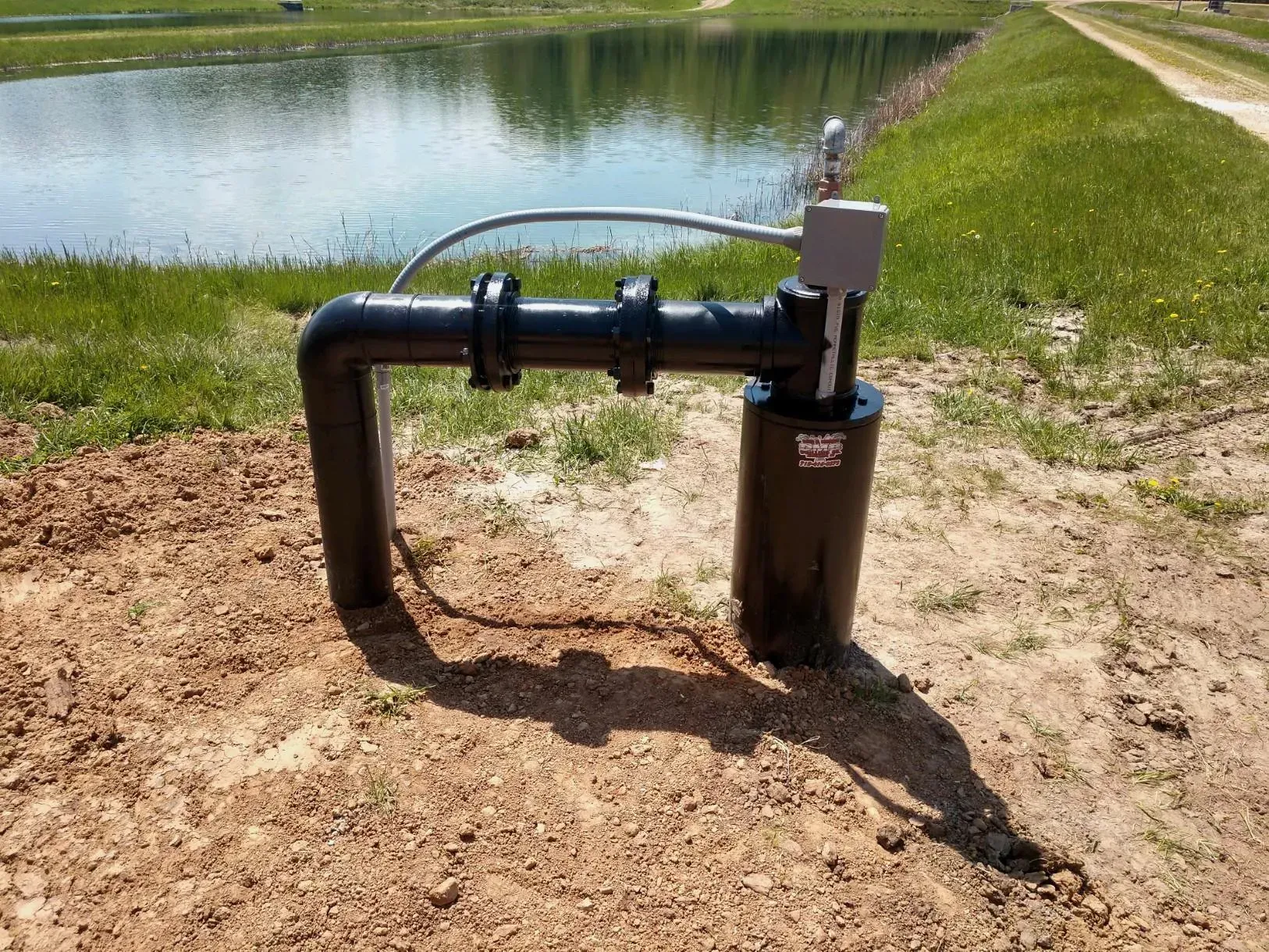 Black pipe system near a pond, likely for water management. Gray metal pipe connects to a control box.