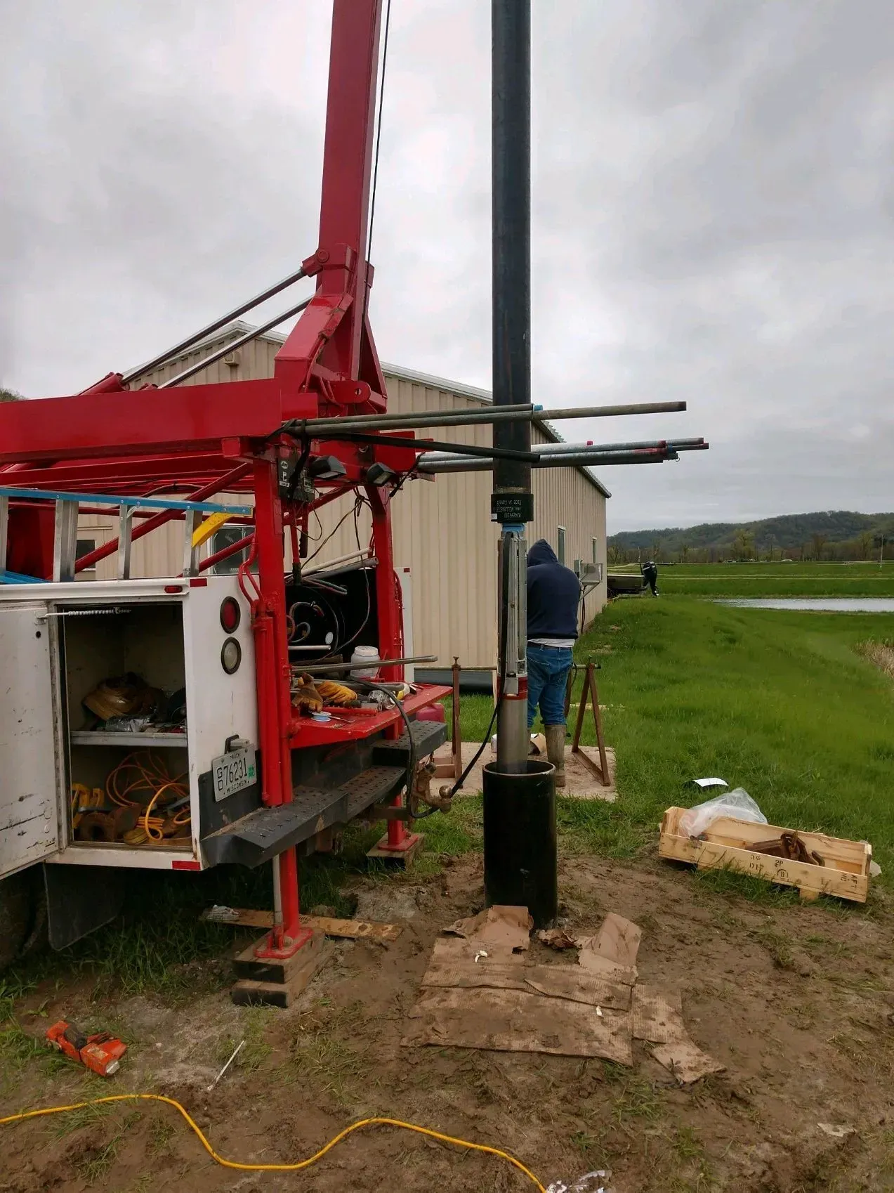 A well-drilling truck with a crane, worker, and well casing near a building and a field.