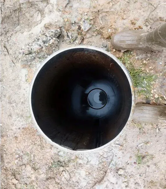 View down into a cylindrical, dark tube set in dirt. A boot is visible at the top right.