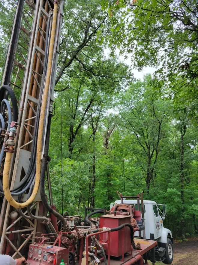 Drilling rig in a forest, cloudy sky. Truck details: red components, white cab. Green trees.
