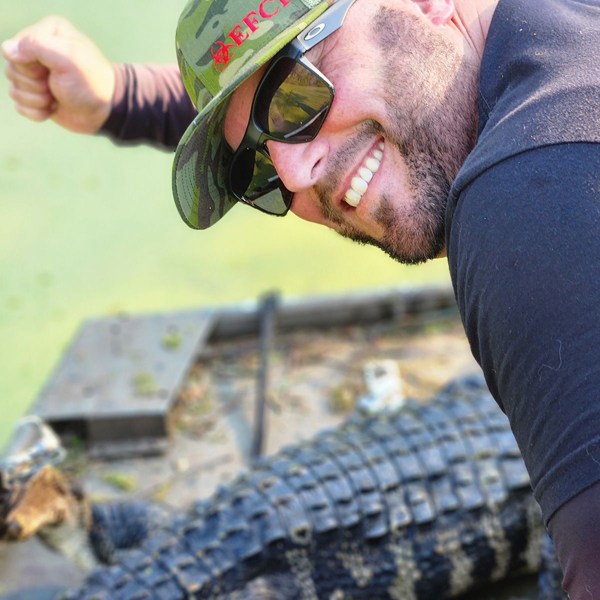 Smiling man in sunglasses and cap posing beside a large alligator or crocodile outdoors
