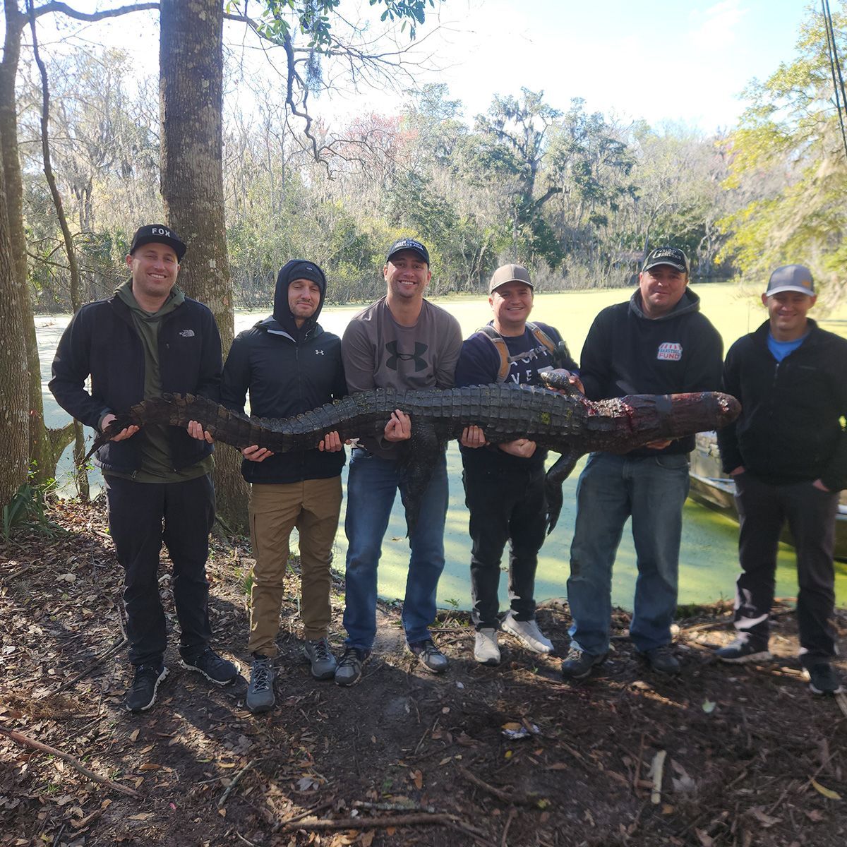 Six people holding a large fish in a wooded outdoor area near a lake