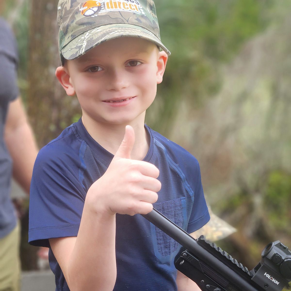 Smiling child in a camo cap giving a thumbs-up while holding a fishing rod outdoors