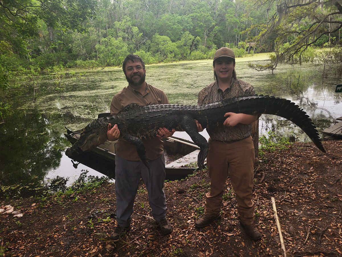 Two people holding a large alligator beside a wooded pond