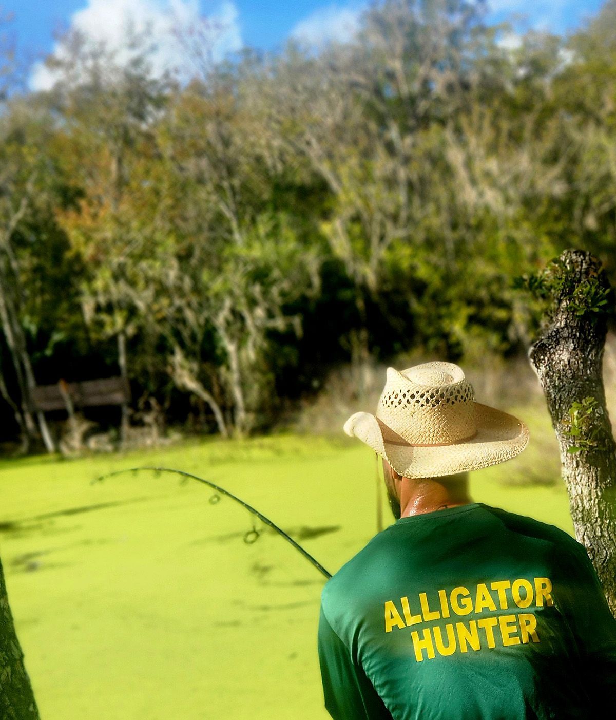 Person in a straw hat and green “Alligator Hunter” shirt holding a fishing rod in a sunny grassy yard.