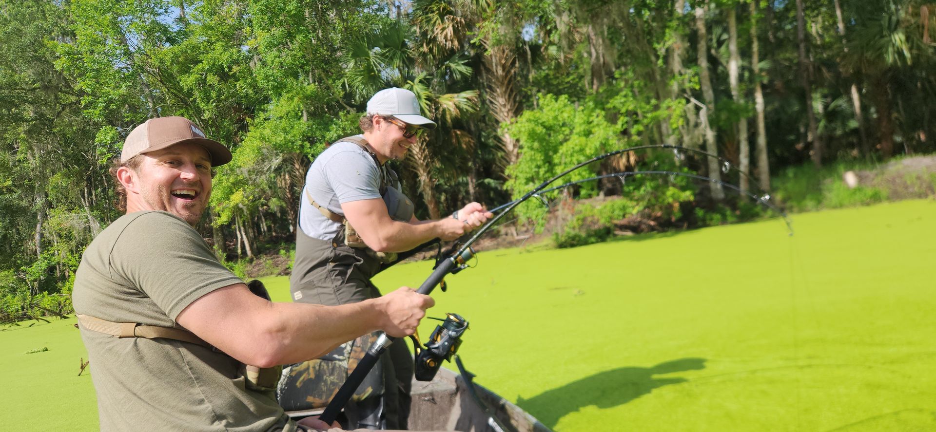 Two men fishing from a boat on a bright green pond near trees