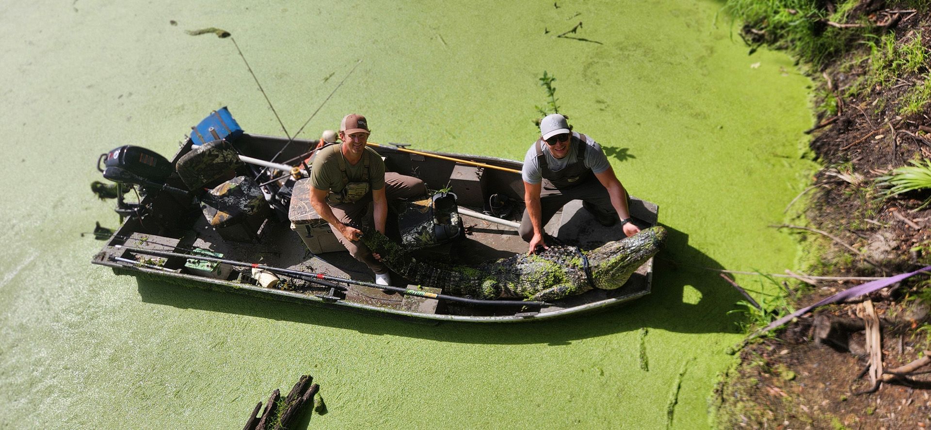 Two people in a small boat floating on bright green algae-covered water near a muddy bank