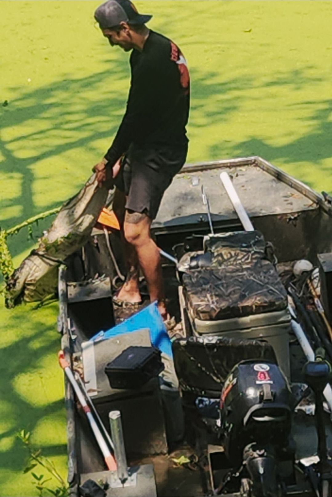 Man standing in a small boat on green water, holding a fish near an outboard motor and fishing gear.