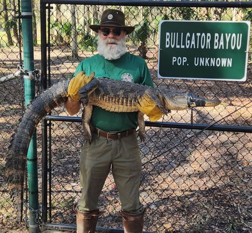 Man holding a large alligator beside a sign reading &ldquo;Bullgator Bayou, Pop. Unknown&rdquo;