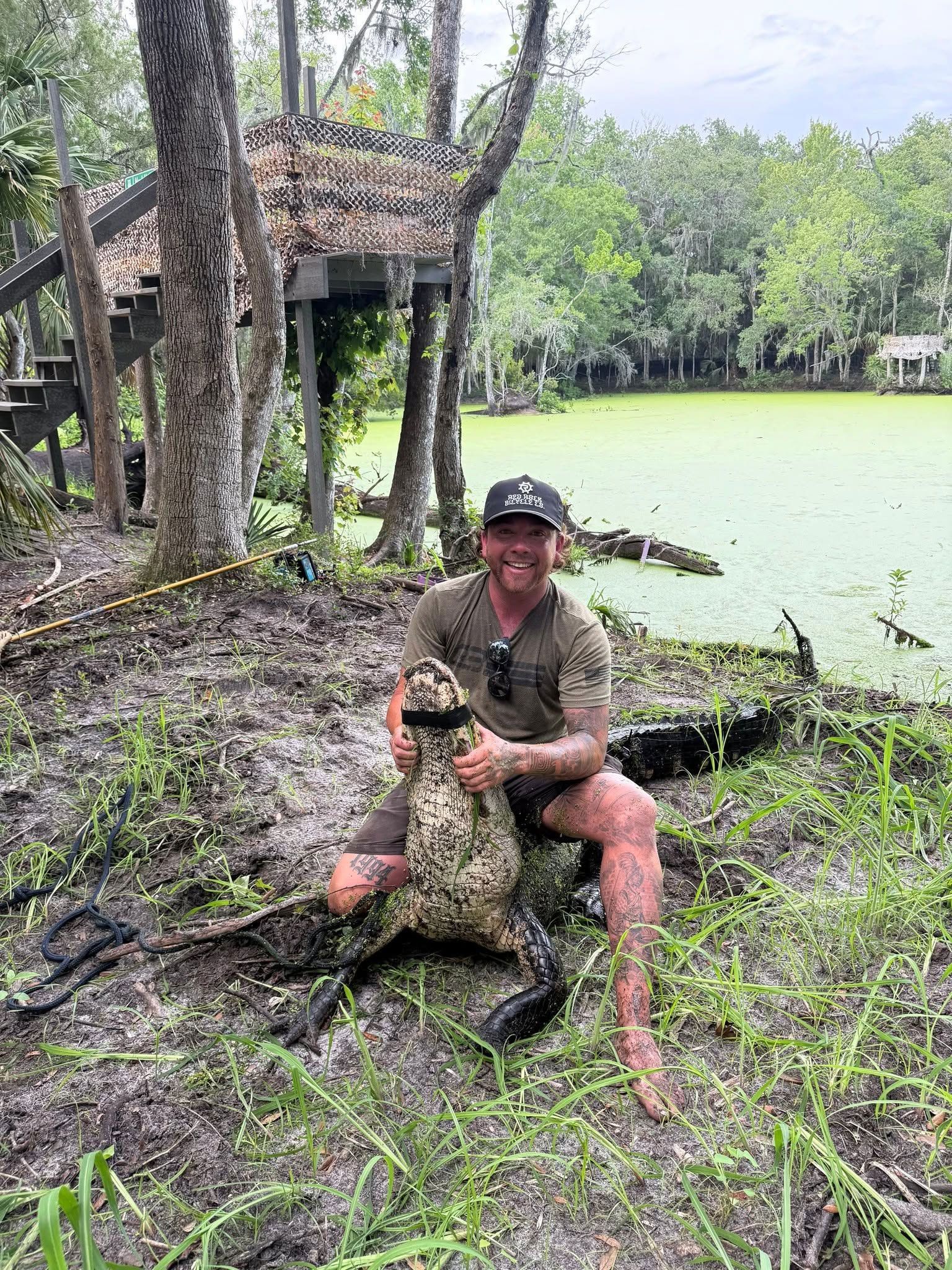 Man kneels beside a large fish on a muddy bank by a green pond, with a small wooden shelter and trees nearby