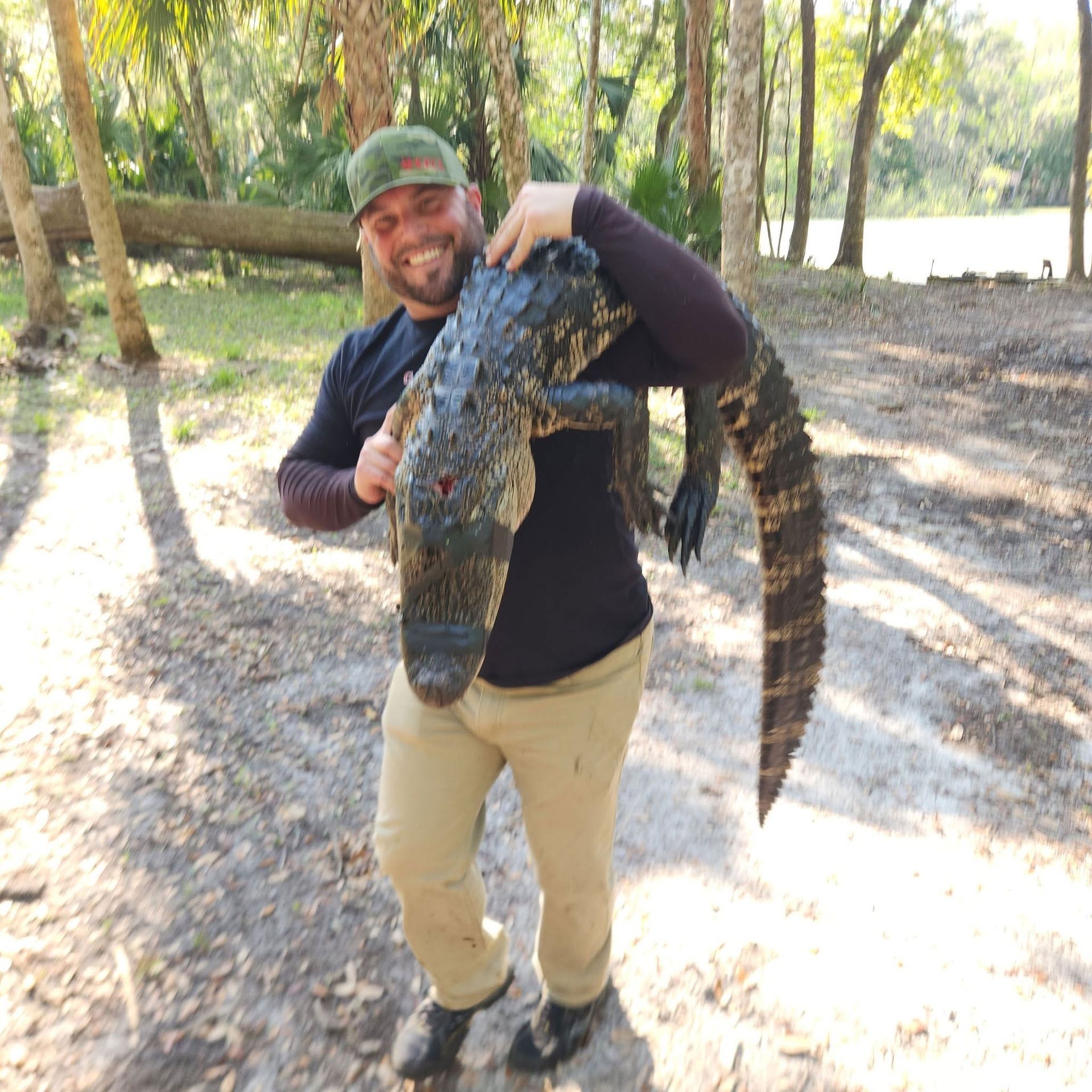 Man holding a large crocodile in a sunny wooded area near water