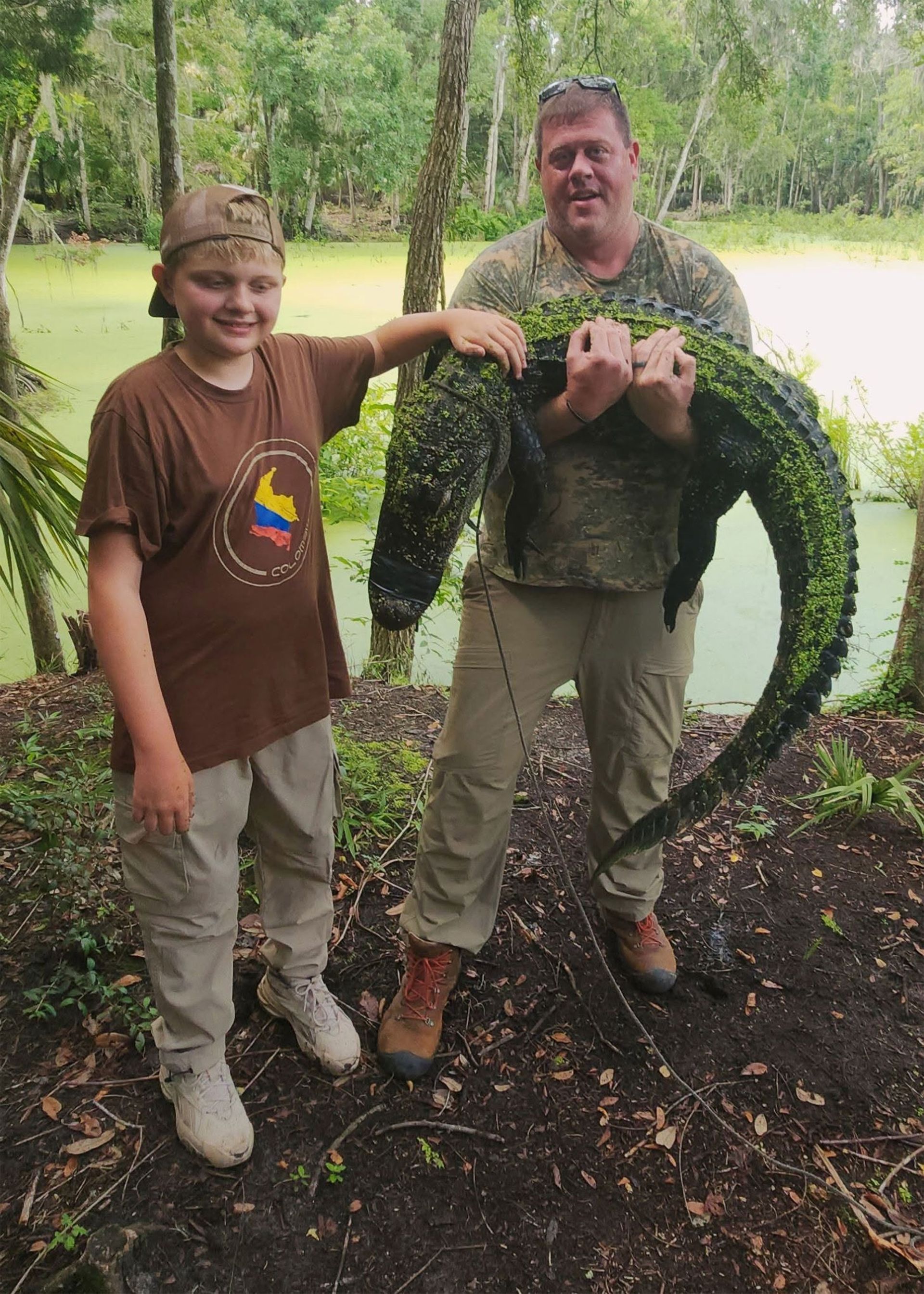 Two people holding a large alligator in a grassy outdoor area