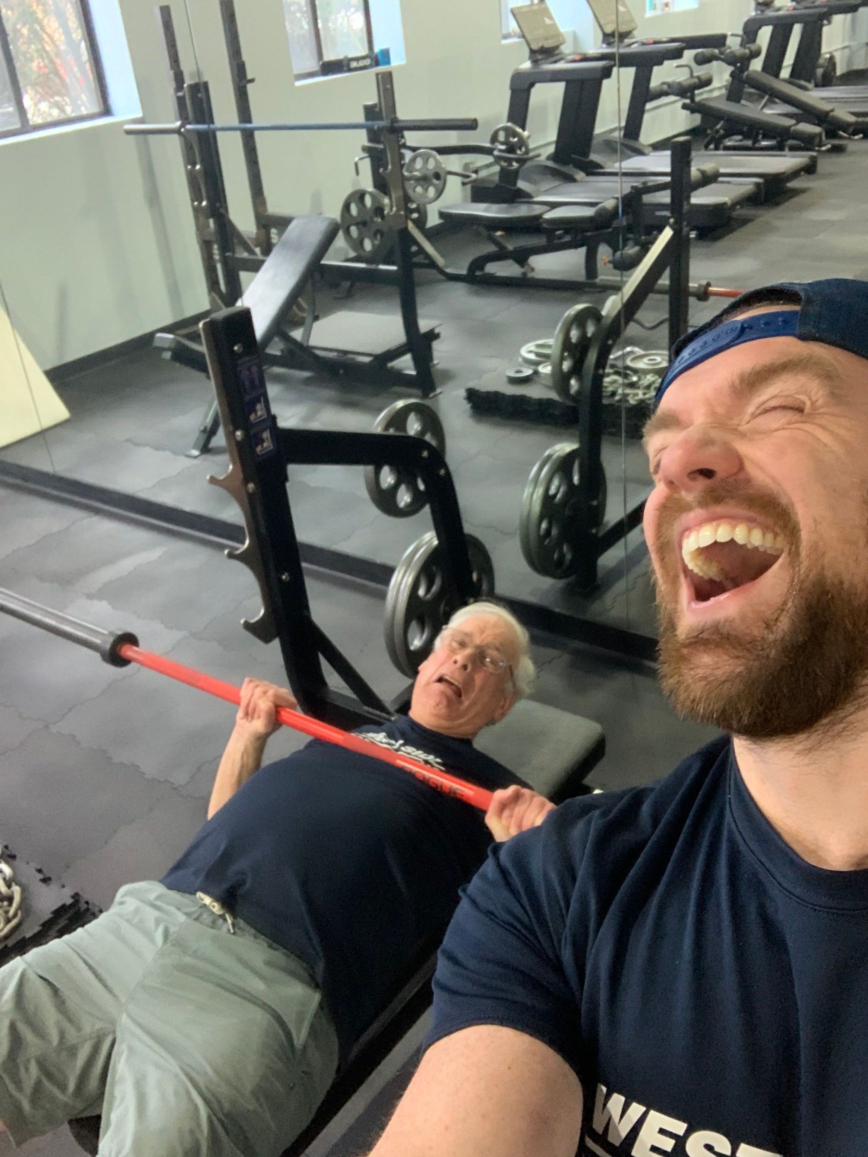 A man is lifting a barbell in a gym while another man sits on a bench.