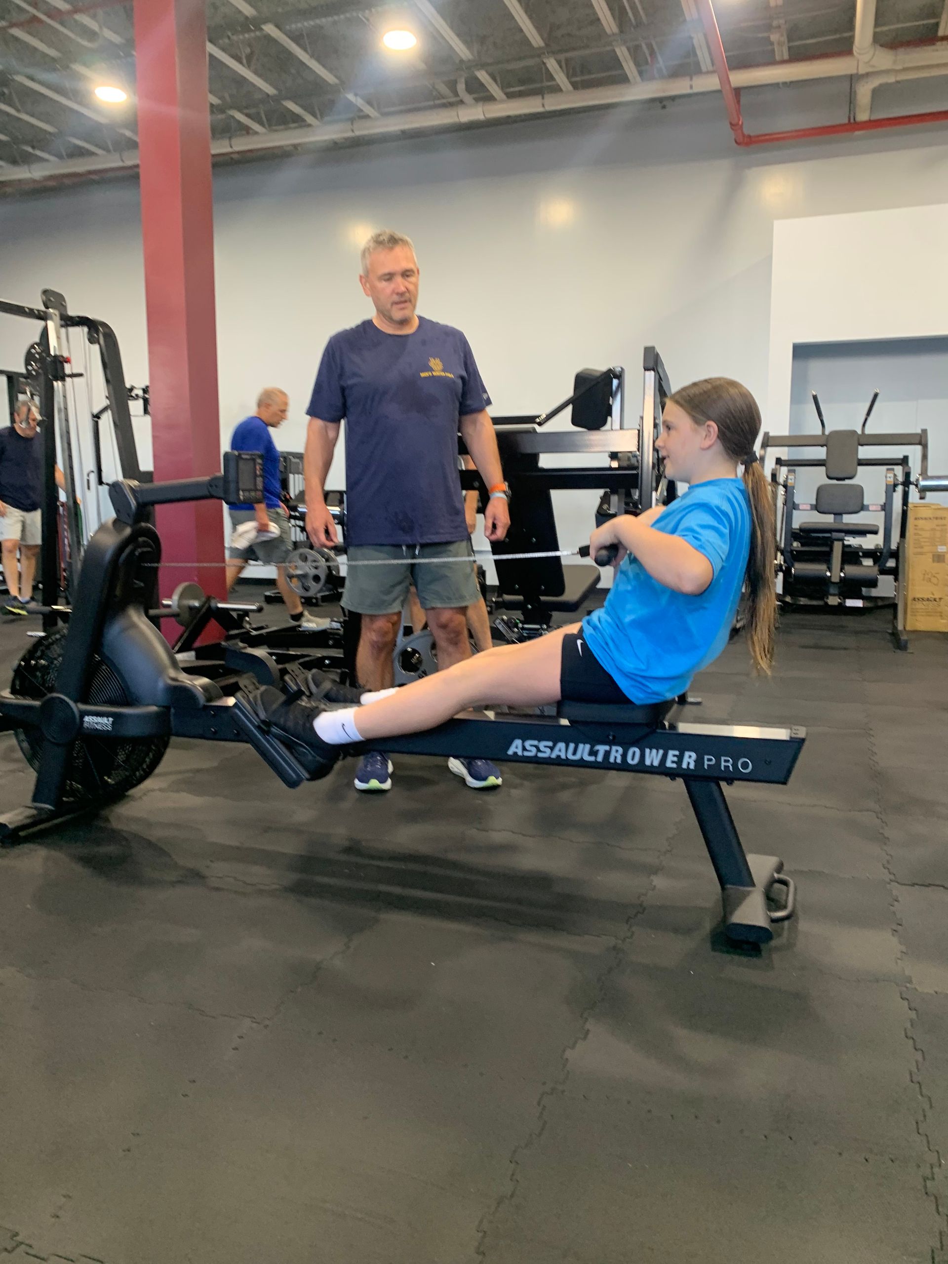 A young girl is using a rowing machine in a gym while a man watches.
