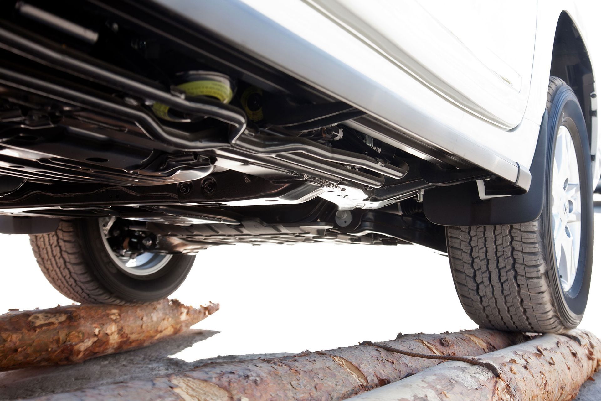 Undercarriage of a white vehicle driving over logs. Tires, mudflaps, and various components are visible.