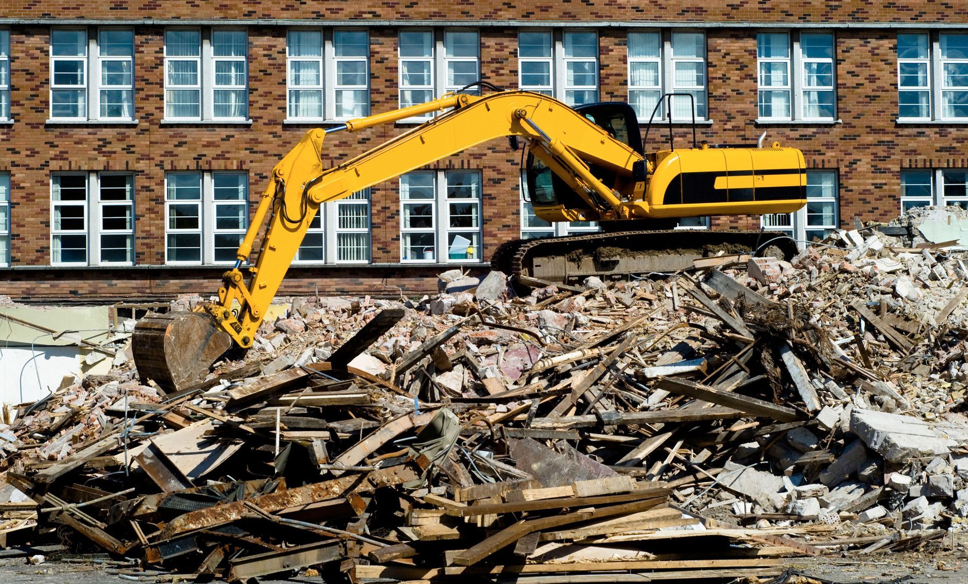 Yellow excavator demolishing building, pile of rubble in front of brick structure.