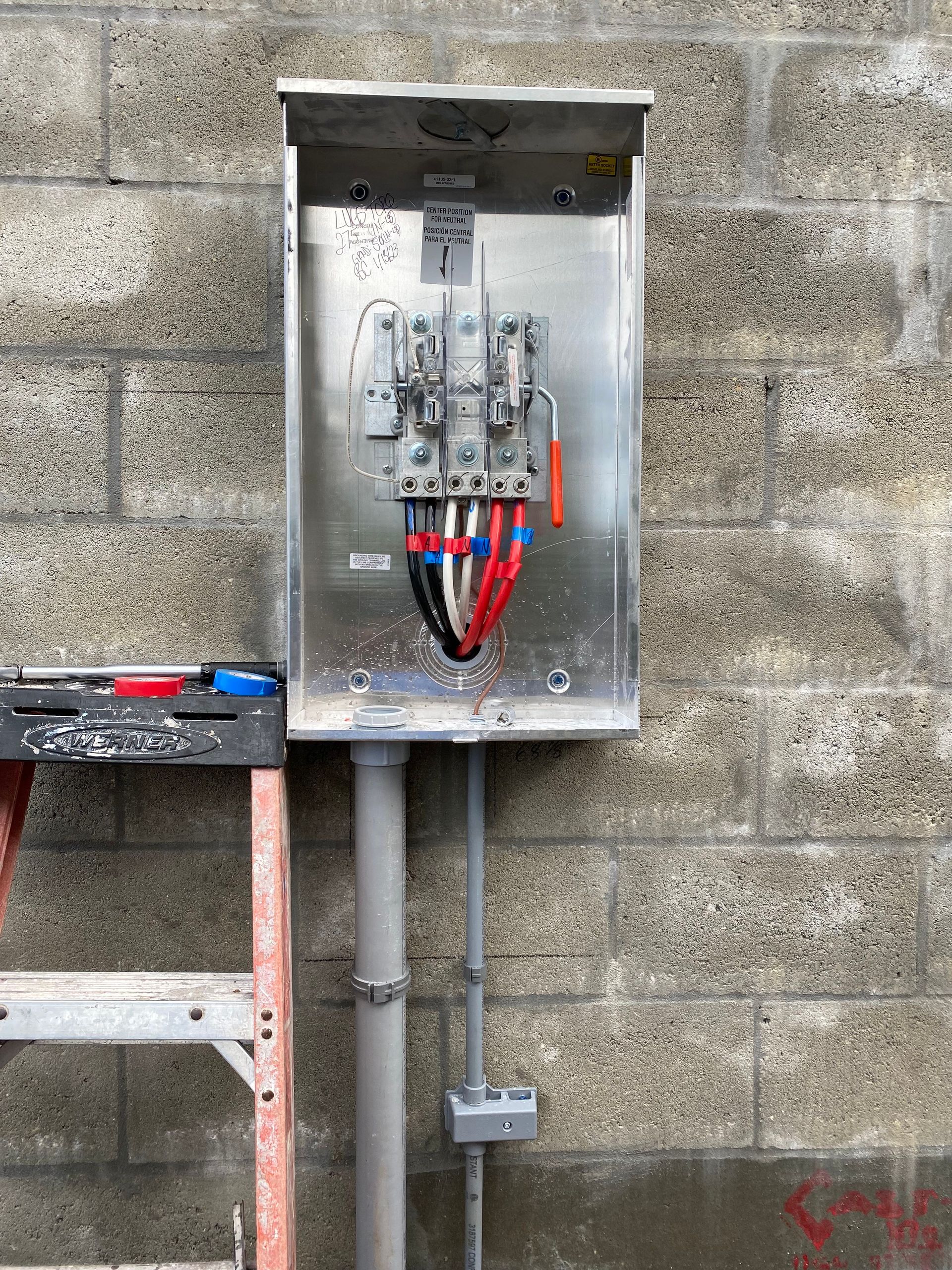 An electrical box is attached to a brick wall next to a ladder
