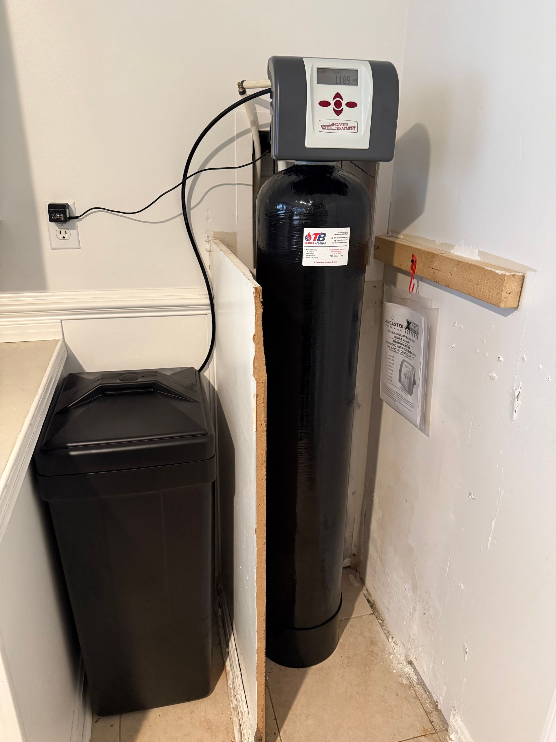 A water softener is sitting in a corner of a room next to a trash can.