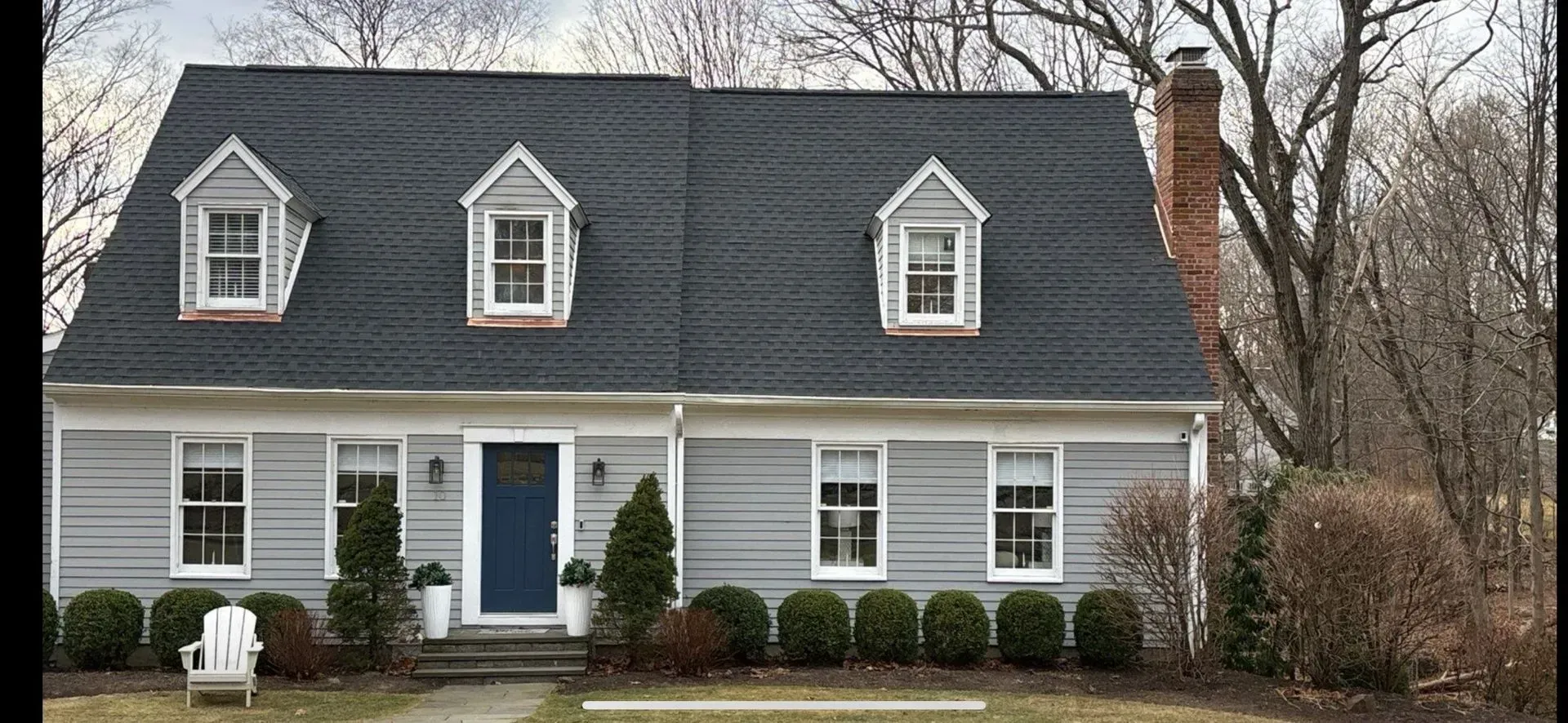 Blue-gray two-story house with dormer windows, white trim, and a brick chimney, surrounded by shrubs and trees