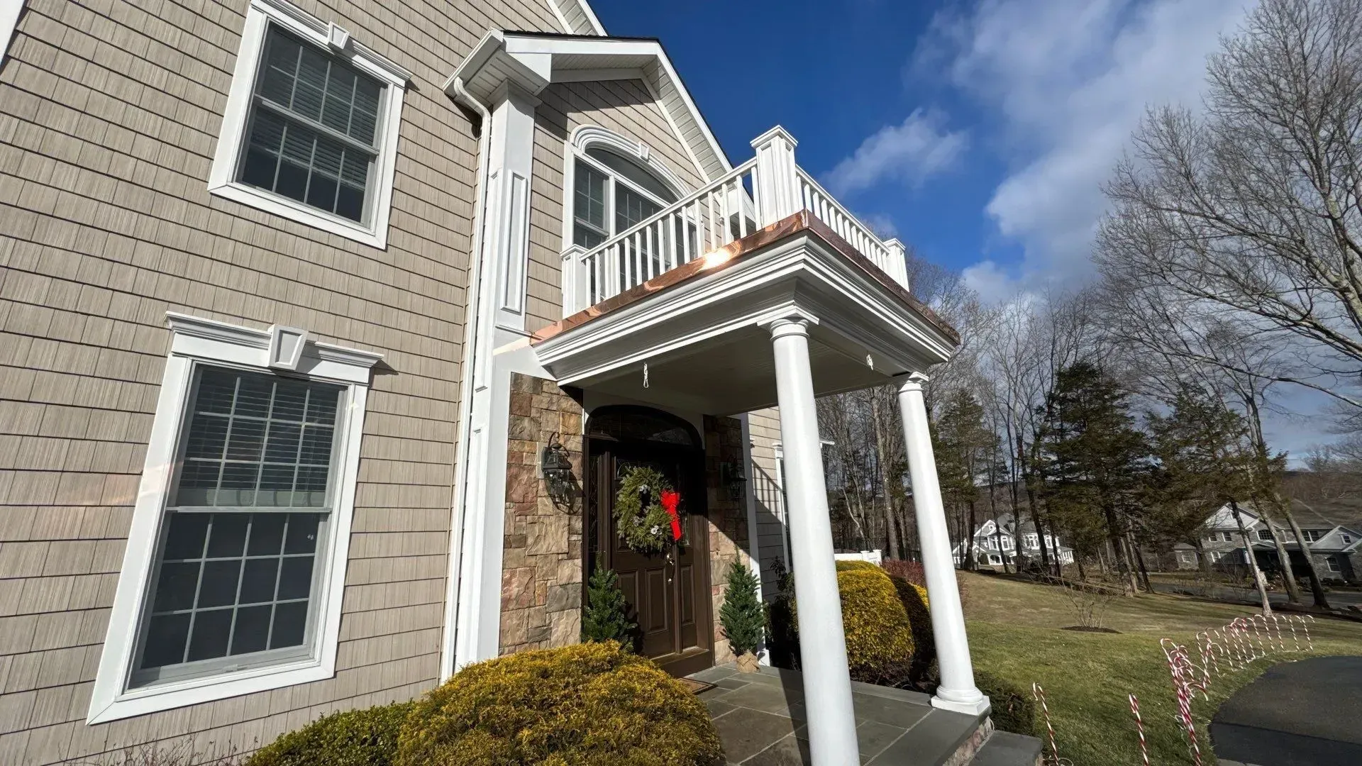 Brick house entrance with white columns, balcony, and wreath on the front door under a blue sky.