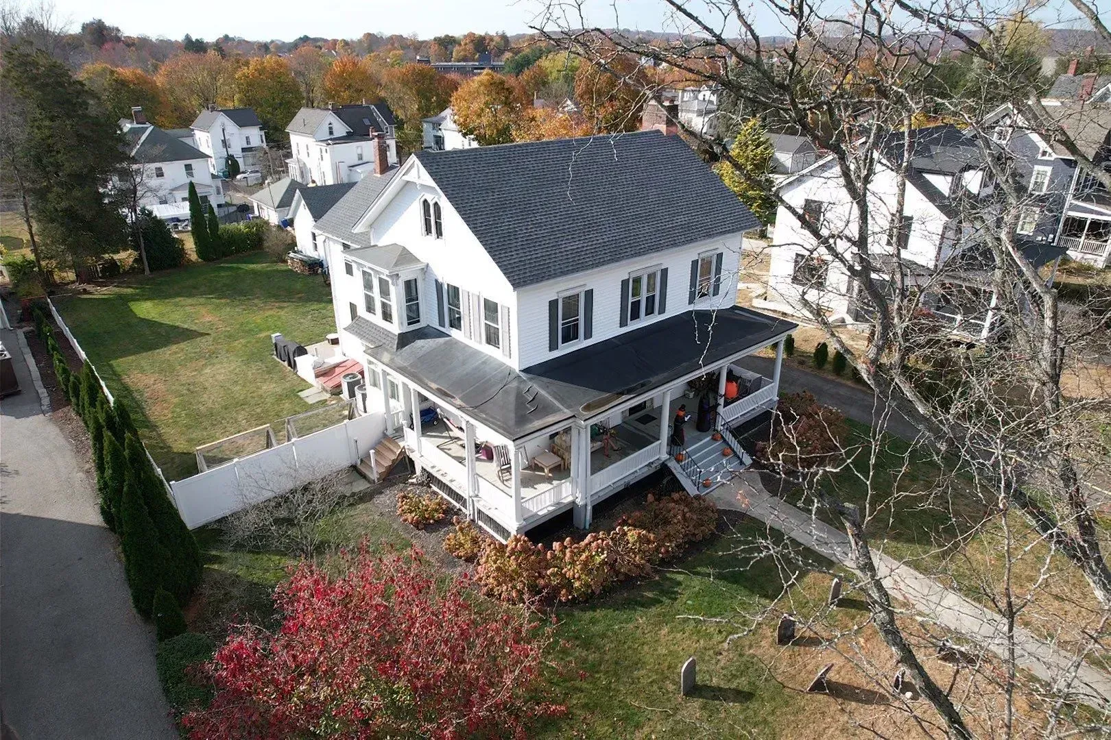 White two-story house with gray roof and wraparound porch in a suburban neighborhood in autumn