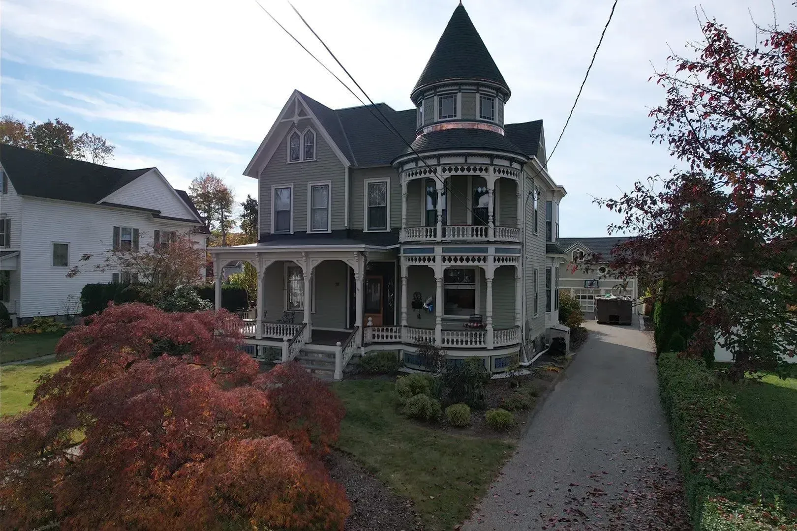 Victorian house with ornate porch and turret beside a driveway, framed by autumn trees