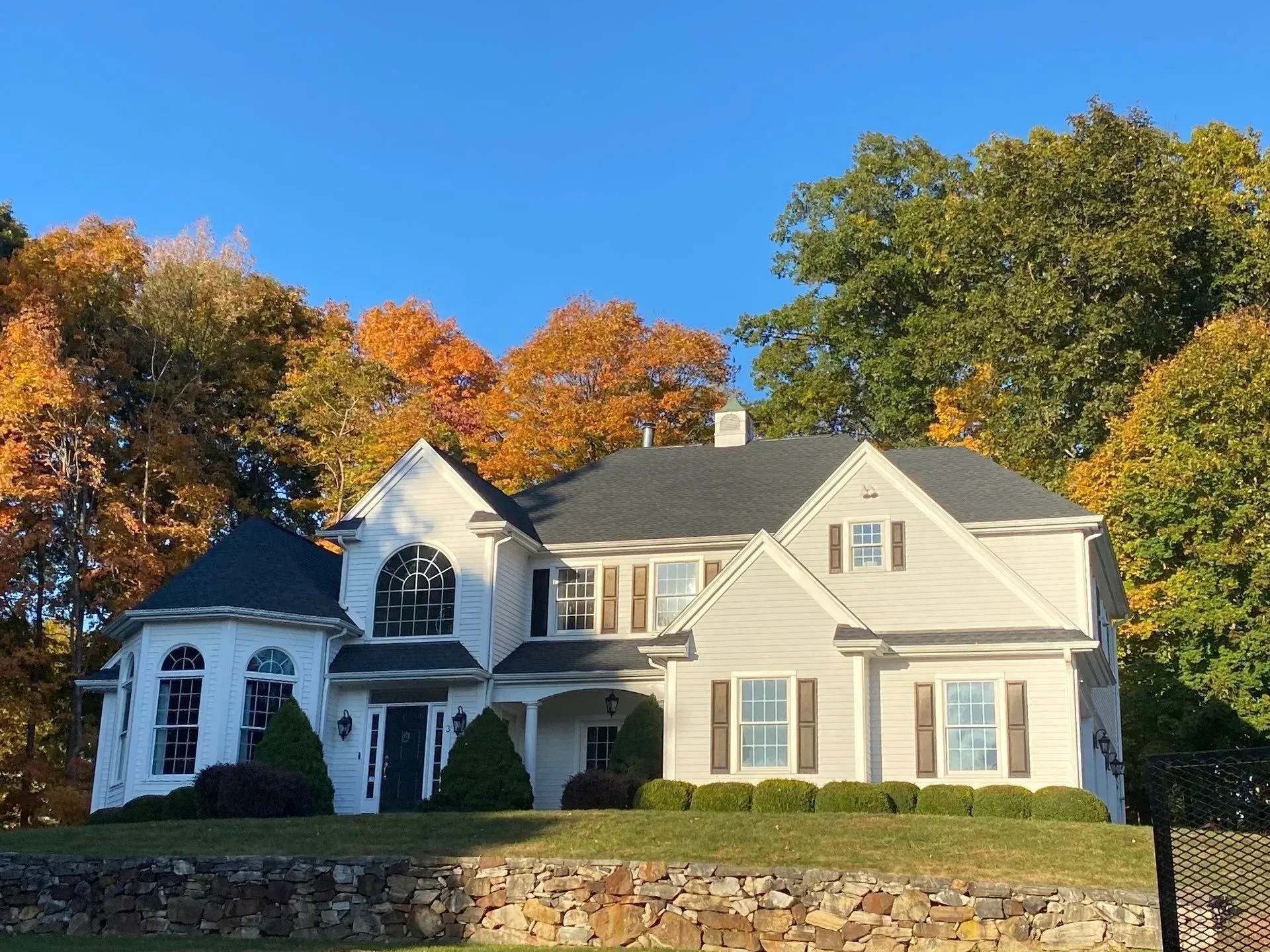 White two-story house with green shutters and autumn trees under a clear blue sky.