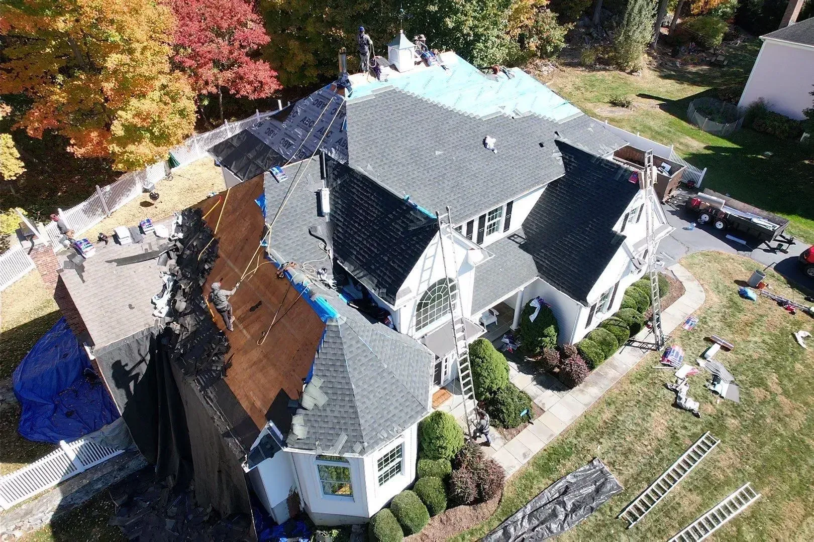 Aerial view of a house with a damaged roof and debris, surrounded by autumn trees and parked vehicles