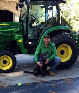 Man kneeling with dog in front of a green and yellow tractor. Exterior setting, smiling.