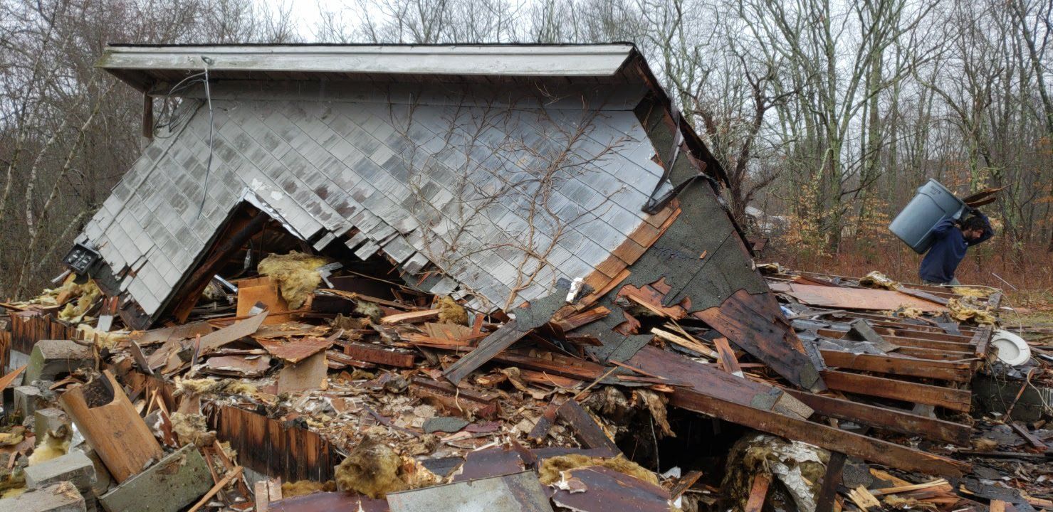 A demolished building with debris and a person carrying a bucket. The building's roof is partially intact.