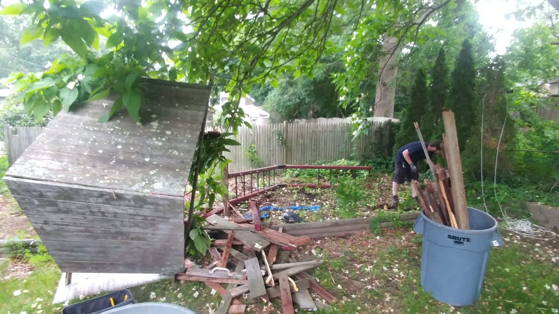 Person clearing debris in a backyard with trees, fence, a large concrete object, and a blue trash can.