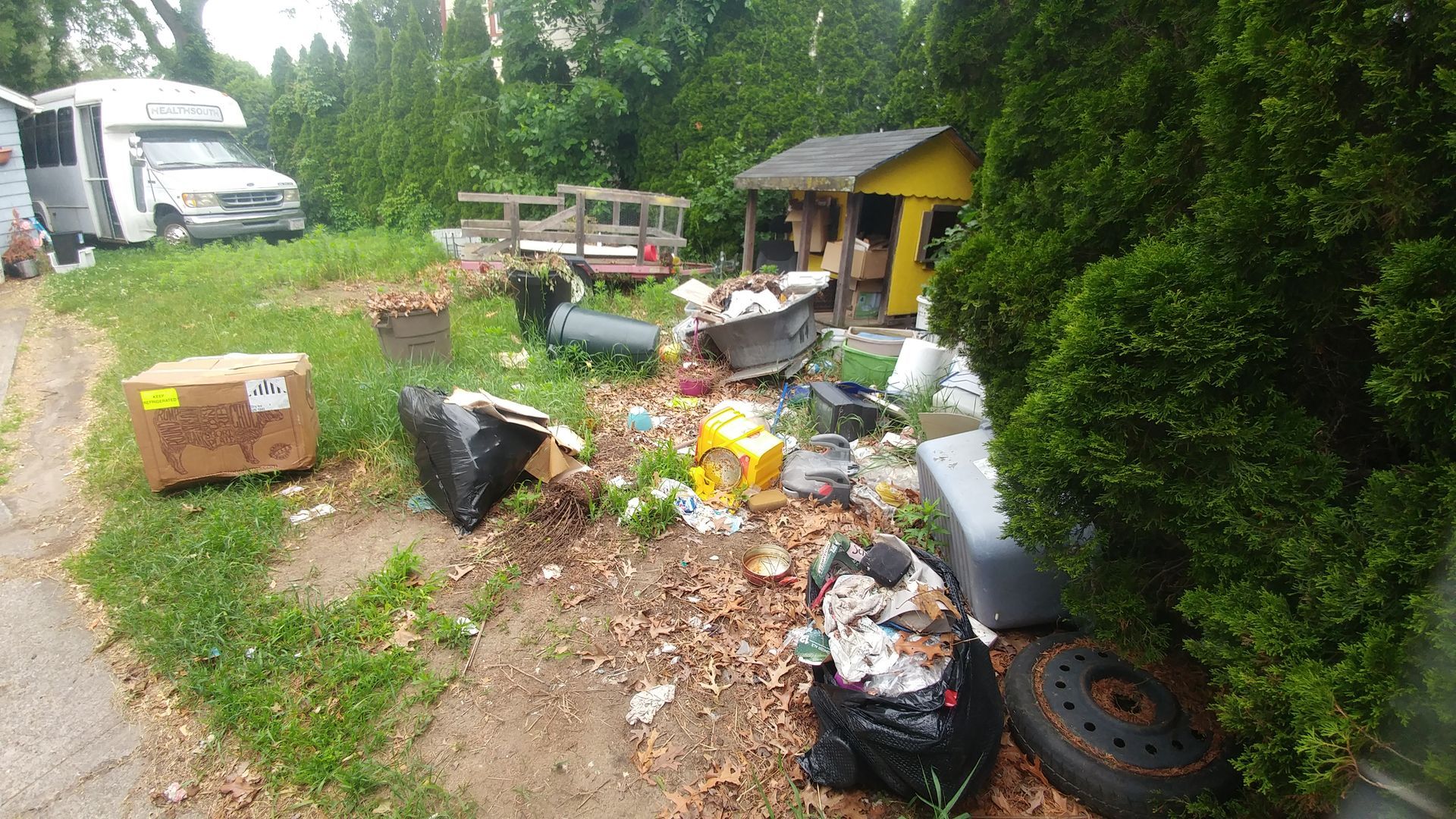 A cluttered yard with trash, a small shed, and a van parked on the side of a road.