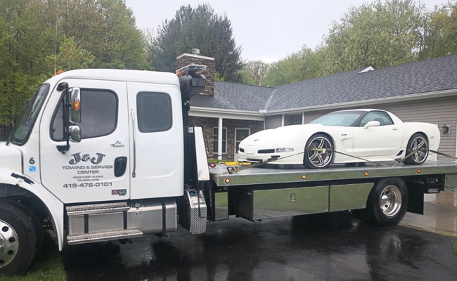 White Corvette being transported on a flatbed tow truck in front of a house. Rain is visible on the ground.