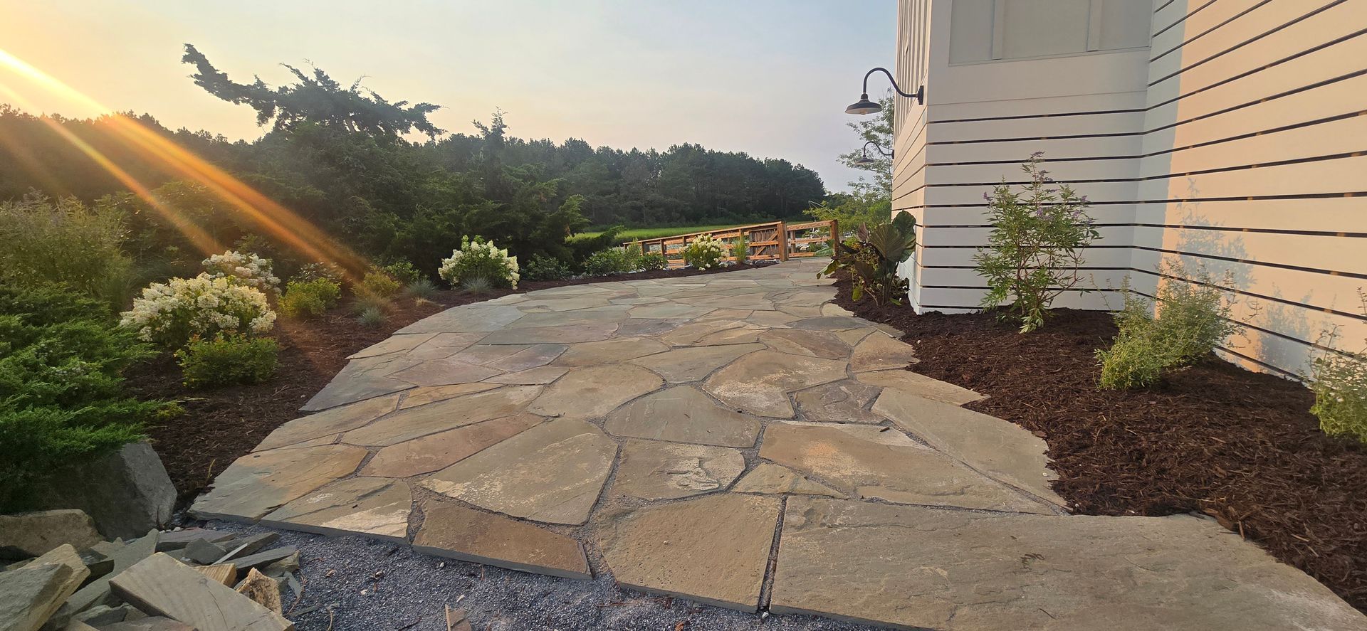 Stone garden path beside house at sunset, with flower beds and a glowing sunbeam.