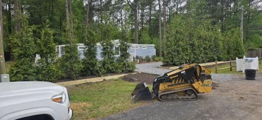 Yellow skid steer parked on a gravel road near a wooded area and white fence posts