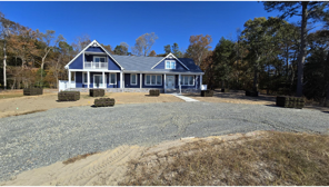 Blue house with white trim, front porch, and gravel driveway on a sunny day. Trees and bushes are in the background.