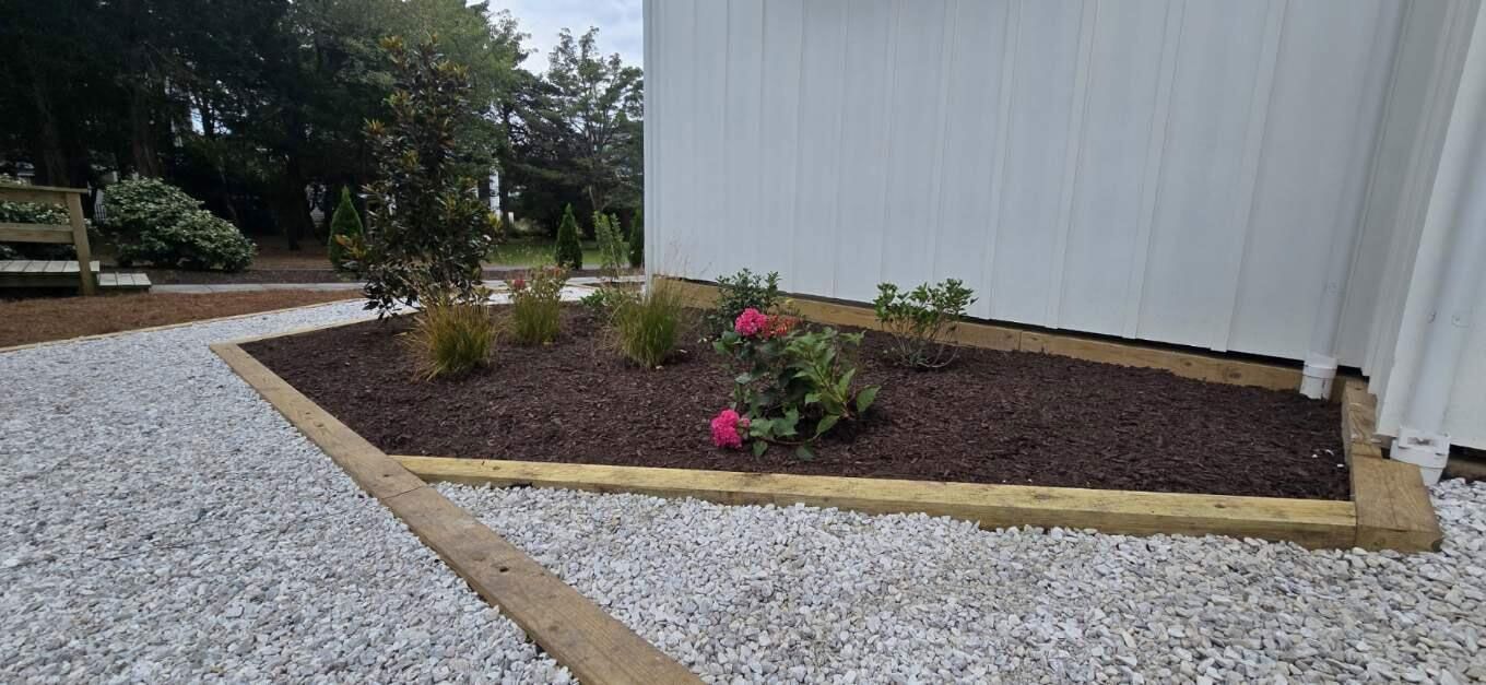 A garden bed with pink flowers and plants, bordered by wood and white gravel, next to a white building.