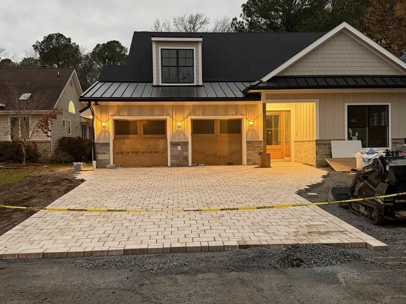 Newly paved driveway in front of a modern house with two garage doors. Brick pavers and a small construction vehicle are visible.