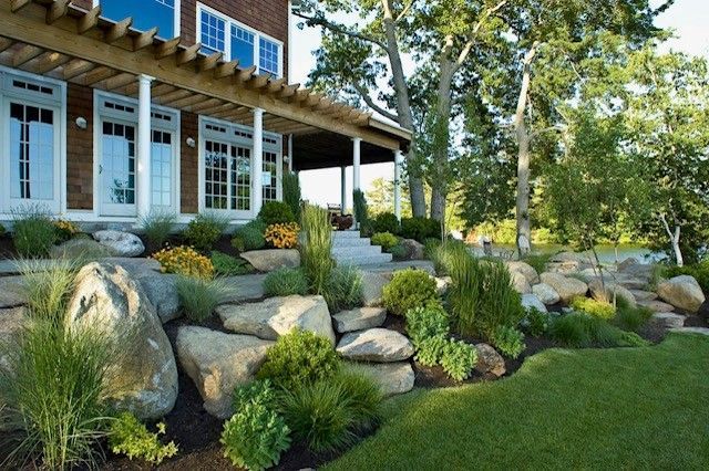 Stone landscaping with shrubs beside a white house and covered porch in a sunny garden