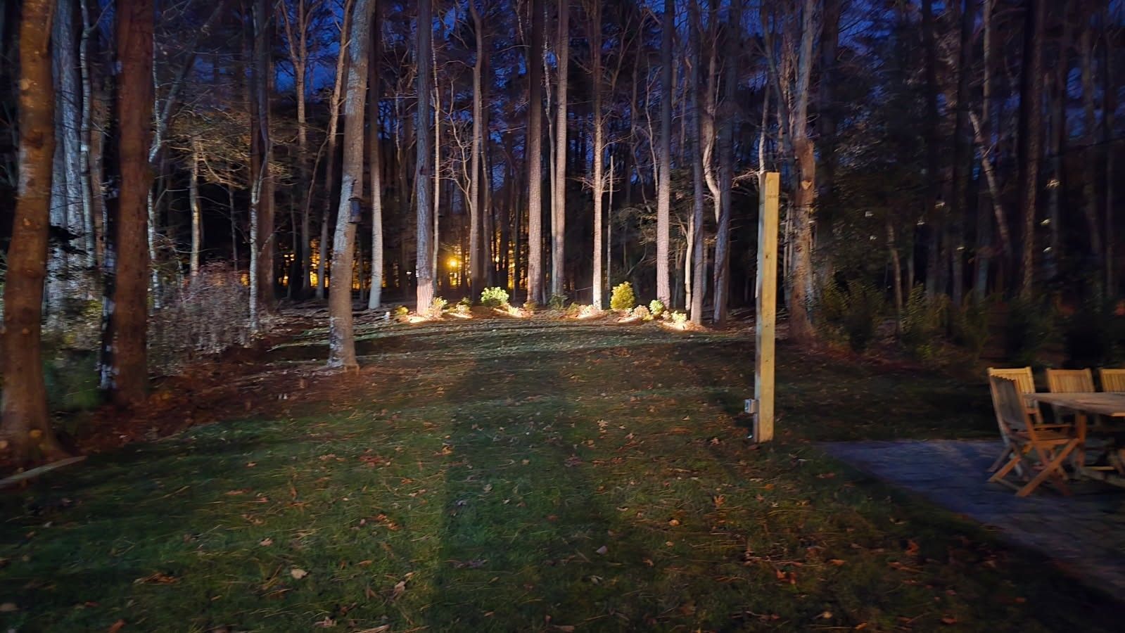 Lawn at dusk, bordered by trees. String lights and outdoor dining furniture are visible.