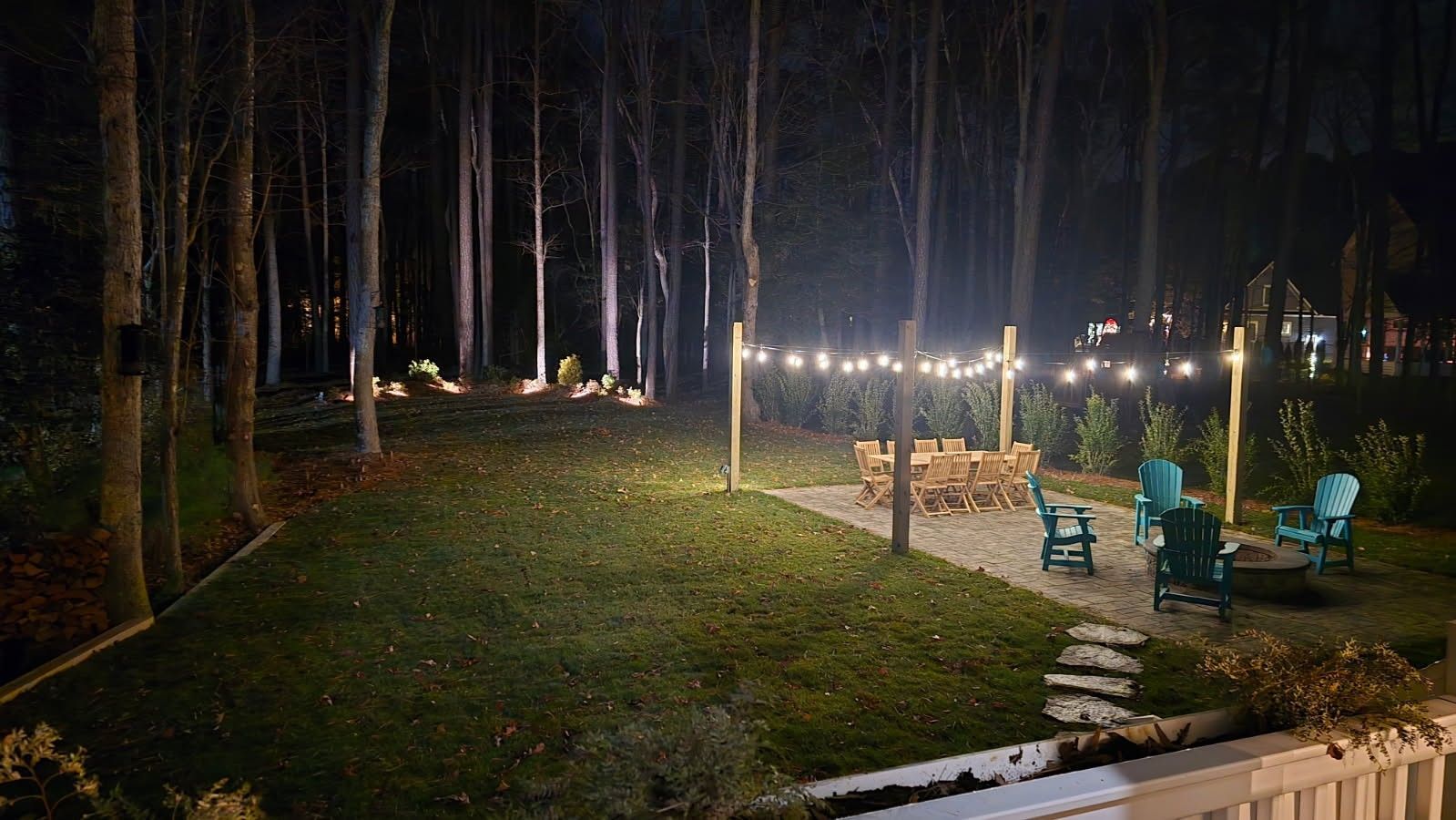 Backyard at night with string lights over a patio with seating. Forest in the background.