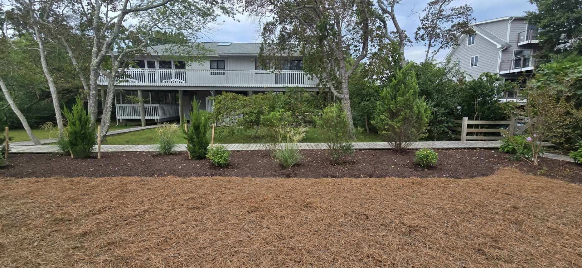 A two-story house with a garden in front. The ground is covered in wood chips, and green bushes are visible.