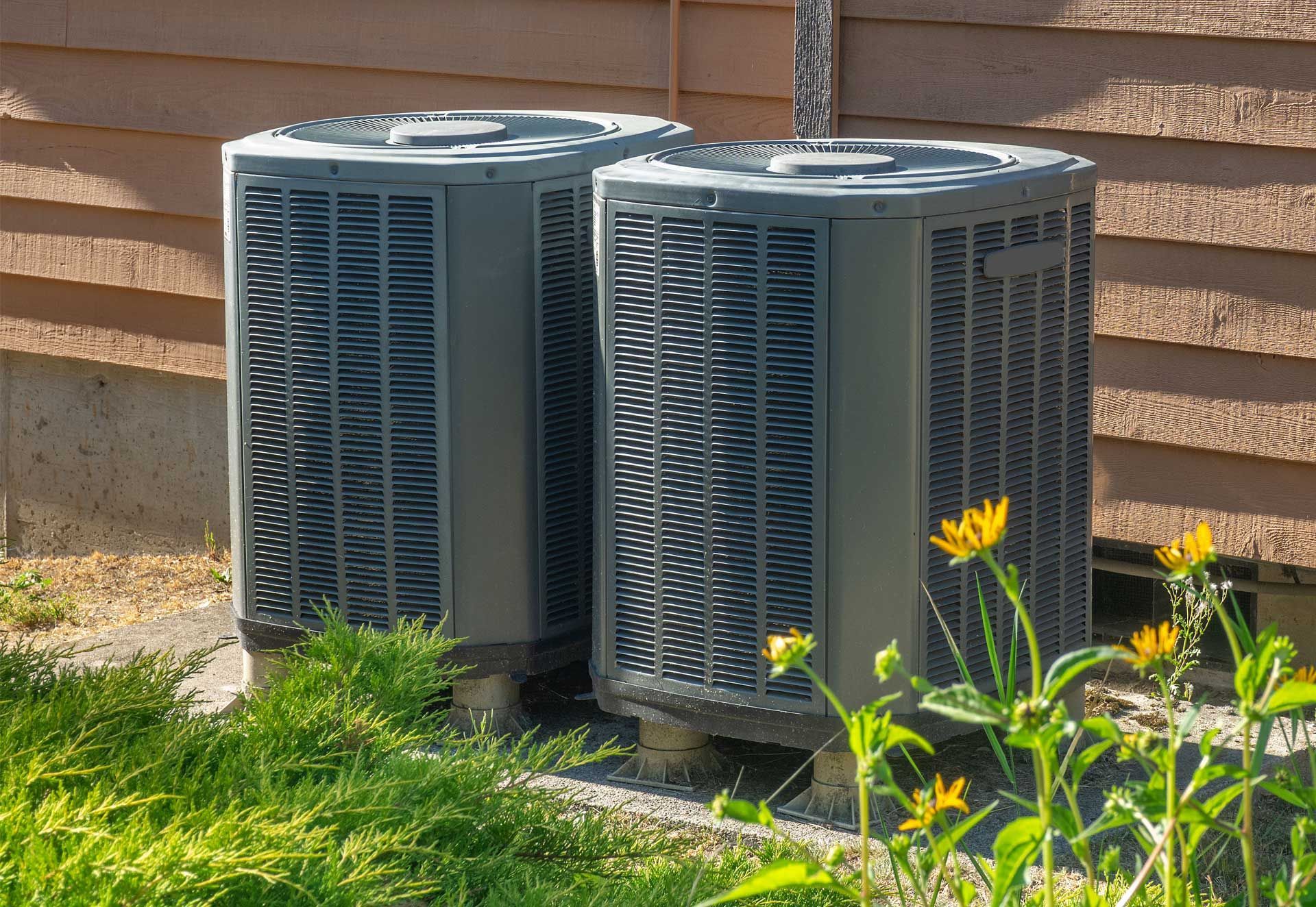 Two outdoor air conditioner units beside a house wall, surrounded by grass and flowers