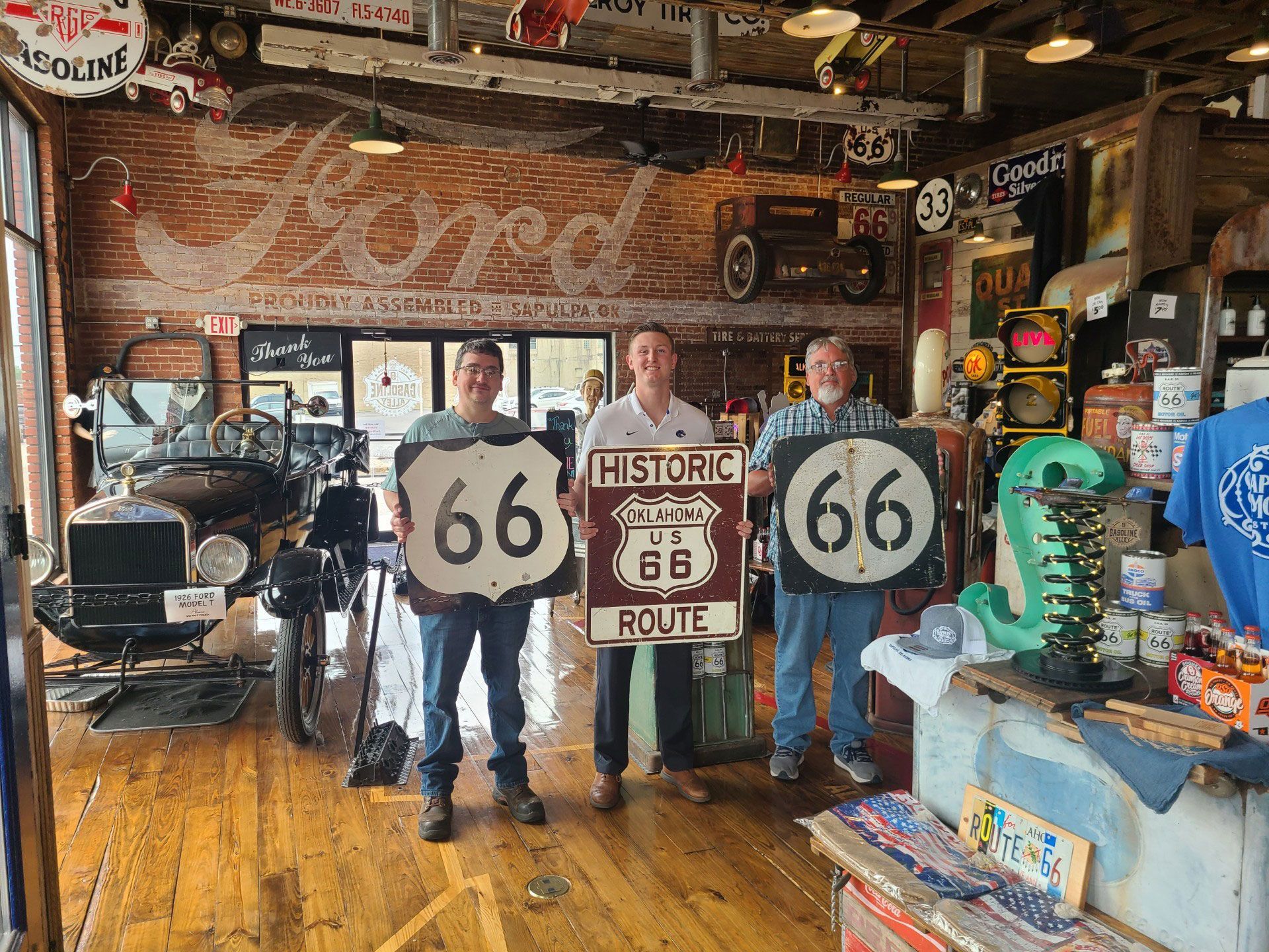 Three people hold Route 66 signs inside a shop with antique cars and Ford sign painted on brick wall.