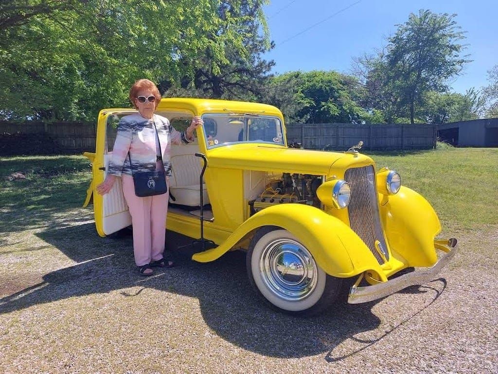 Woman in pink pantsuit stands beside a bright yellow vintage car with open door. Outdoors on a sunny day.