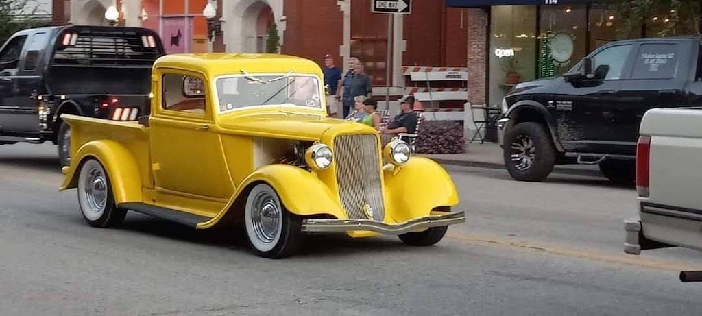 Yellow vintage pickup truck on a city street. Other vehicles and people in the background.
