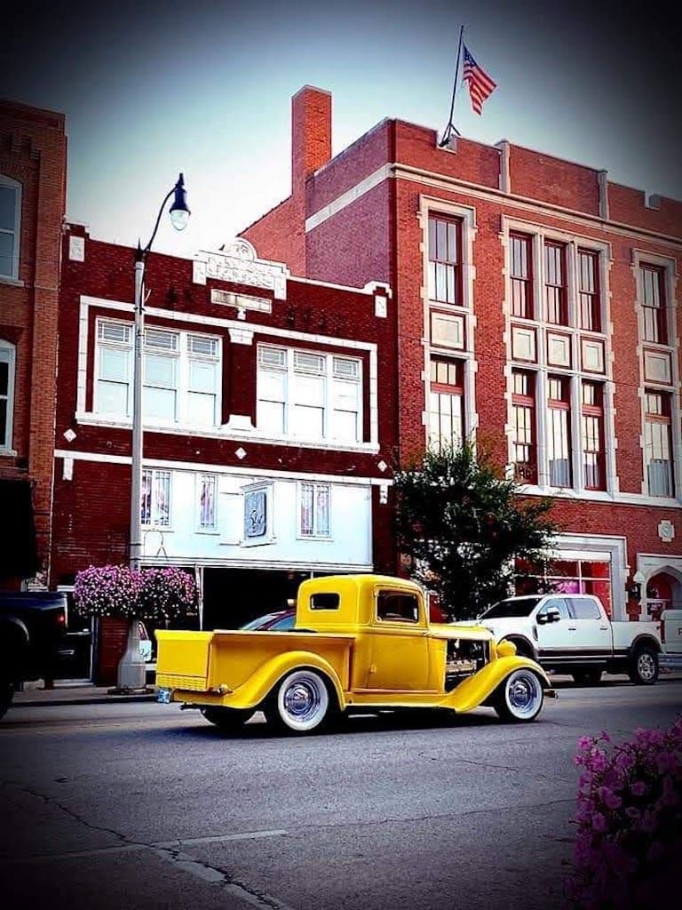 Yellow classic pickup truck driving on street in front of red brick buildings with American flag.
