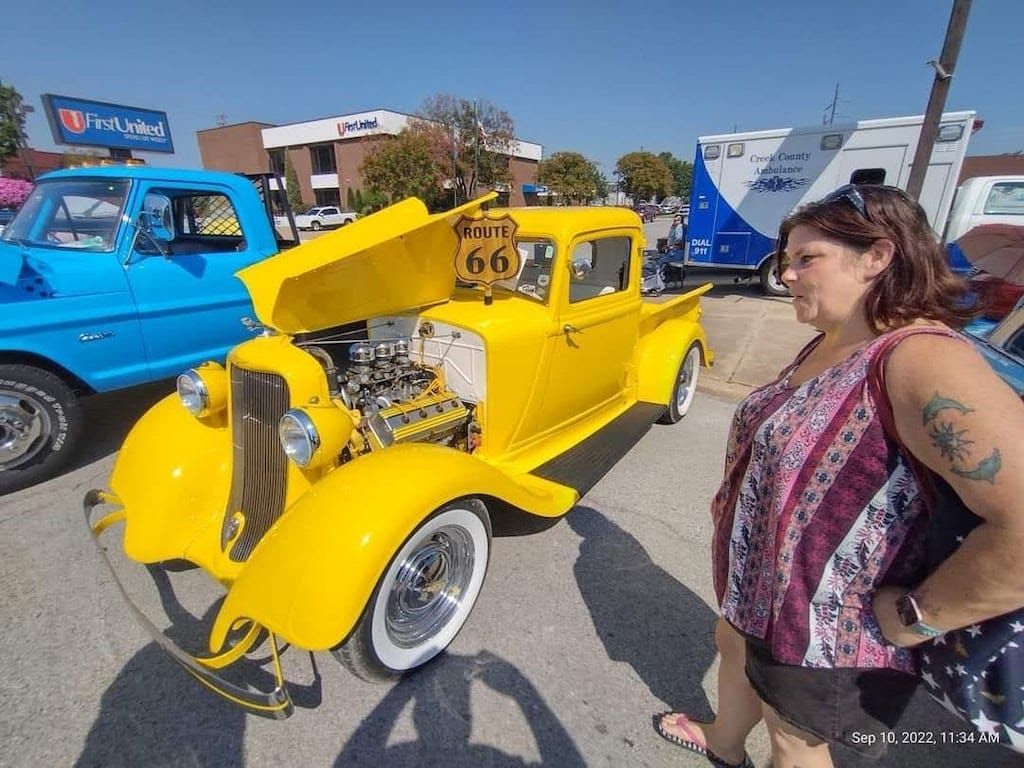 Yellow classic car with Route 66 sign, woman looking at the vehicle at an outdoor car show.