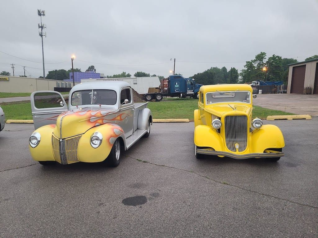 Two yellow vintage cars parked outside on a cloudy day. One with flames on the hood, the other without.